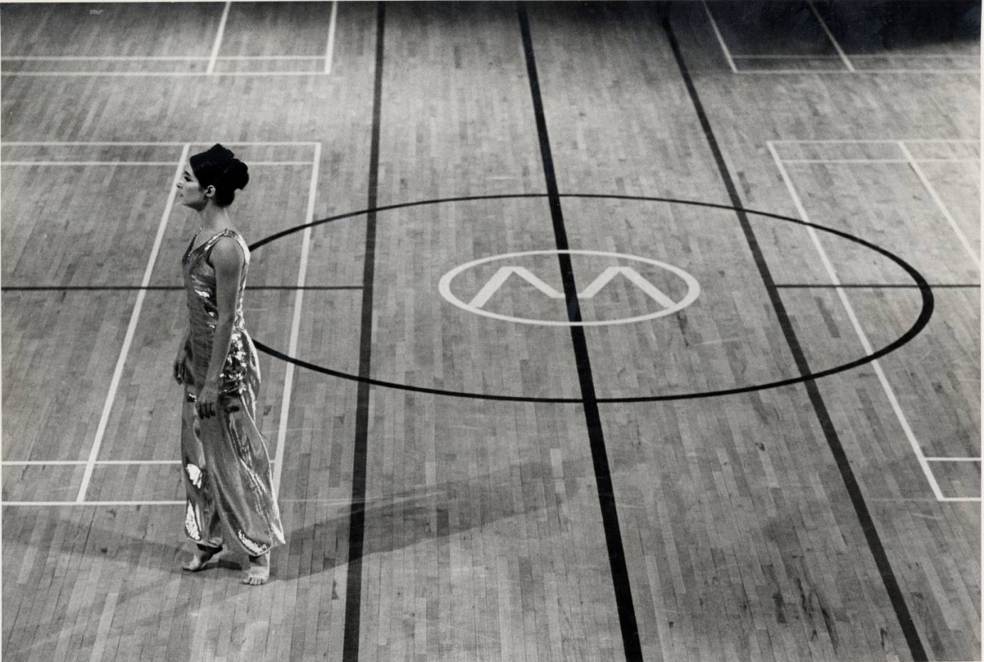 A woman stands in relve in side profile on basketball court. She's barefoot and wears an iridescent costume.